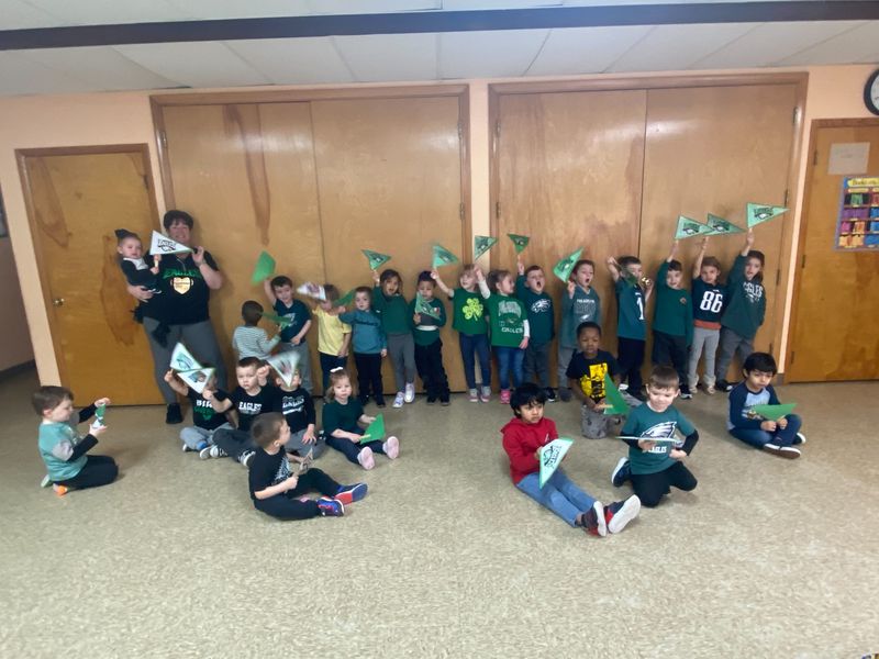 Children in green shirts holding paper flags for St. Patrick's Day with their teacher